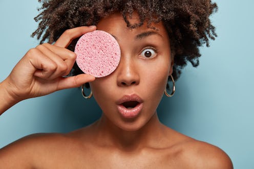 Surprised dark skinned woman applies face foundation, holds pink sponge on eye, has spa procedure an...