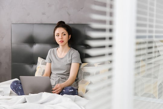 A beautiful young Asian woman in home clothes and glasses is sitting on the gray bed and holding a l...