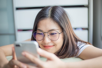Happy young glasses smiling asian women using smartphone on cozy bed texting messages