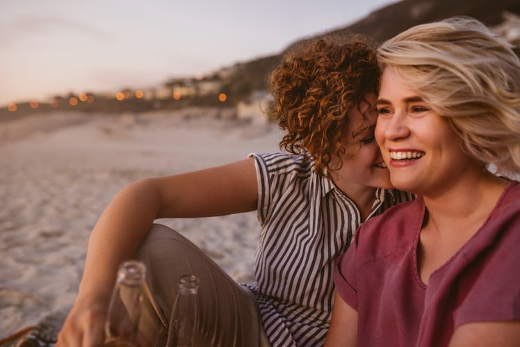 Laughing young lesbian couple talking and having drinks while sitting together on a sandy beach watc...
