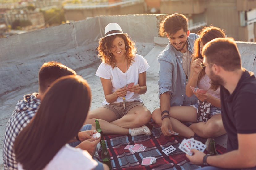 Group of young people sitting on a picnic blanket, having fun while playing cards on the building ro...