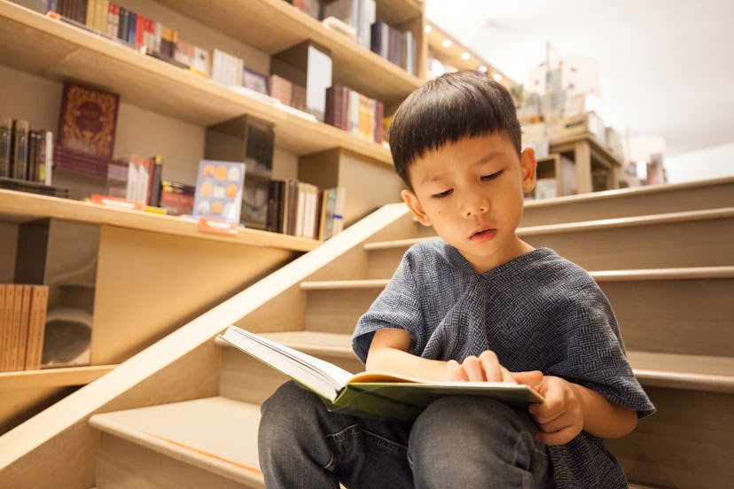Portrait shot of an Asian pre-school boy sitting on the stairs, reading book in the library paying f...