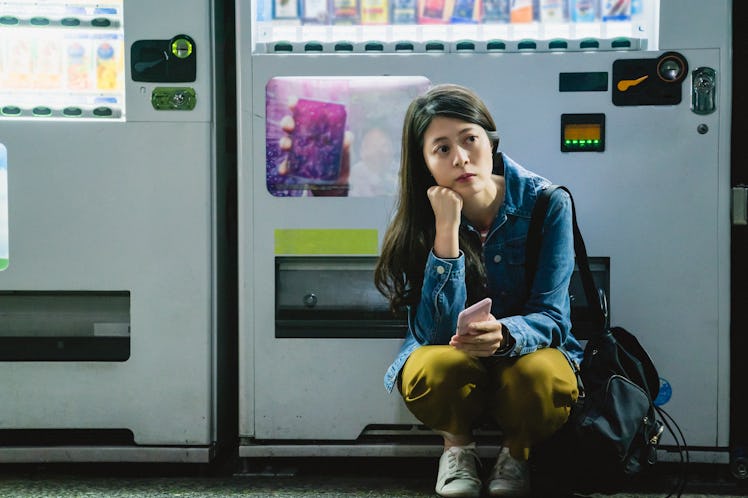 asian girl squatting down and holding her phone to wait for something with a confused thinking and t...