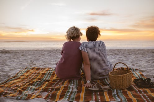 Rearview of a content young lesbian couple sitting together on a blanket at the beach watching a rom...