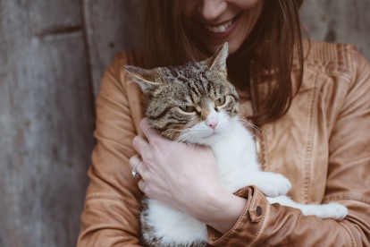 A brunette woman in a light brown leather jacket smiles while holding her cat. 