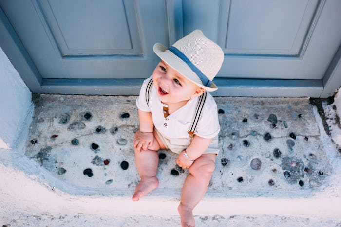Beautiful baby sitting in front of a gray door in Santorini, Greece, wearing a hat