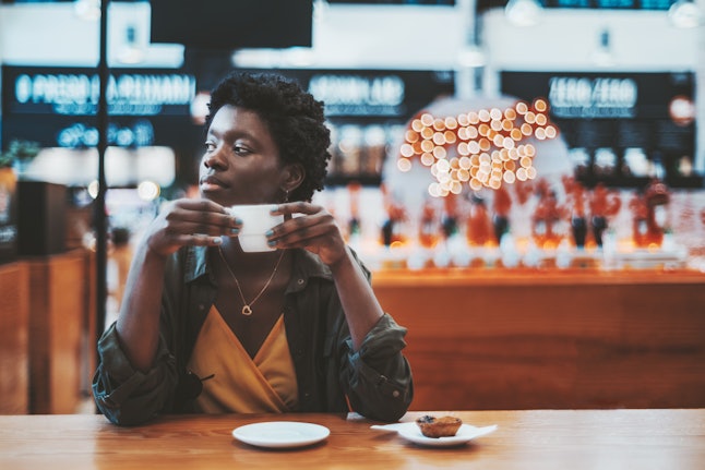 Pensive young African female is having breakfast in a cafe indoors of a mall or a food market and pensively looking aside while holding a cup of delicious coffee; a copy space place on the right