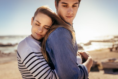 Smiling woman hugging her boyfriend from behind with eyes closed standing on a beach. Couple spendin...