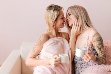 Lesbian couple sitting on pink couch drinking hot coffee kissing each other. Morning same-sex couple...