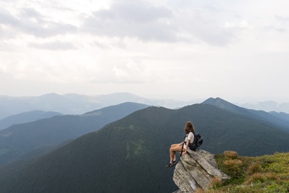 Young female hiker sits on rock in gorgeous mountain area and looks in endless horizon. Woman with t...