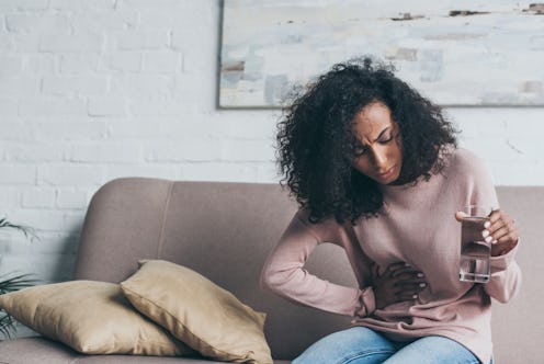 unhappy african american woman holding glass of water while sitting on sofa and suffering from stoma...