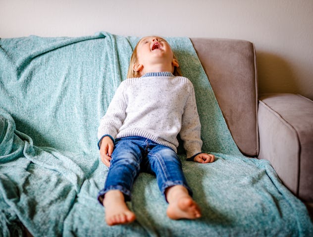 Cute toddler boy screams and cries on couch. Child is wearing blue jeans and white sweater while sit...