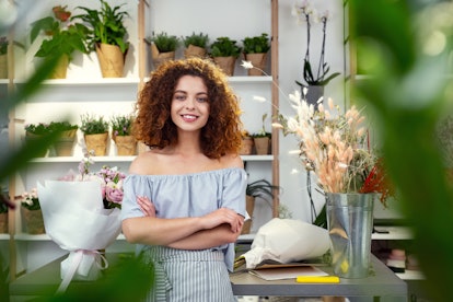 Friendly smile. Joyful confident woman looking at you while standing cross handed in the flower shop