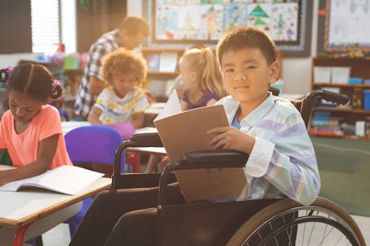 Front view of schoolboy looking at camera while sitting at desk in school against school kids in ba...