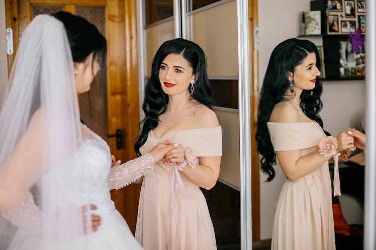 Bridesmaids helping slender bride lacing her wedding white dress, buttoning on delicate lace pattern...