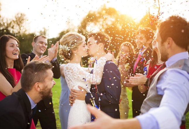 Full length portrait of newlywed couple and their friends at the wedding party showered with confetti in green sunny park
