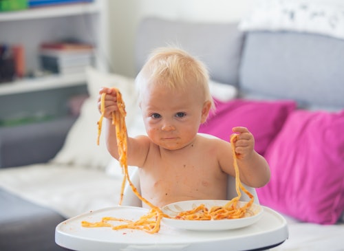 Adorable little baby one year old eating pasta indoor. Funny toddler child with spaghetti. Cute kid ...
