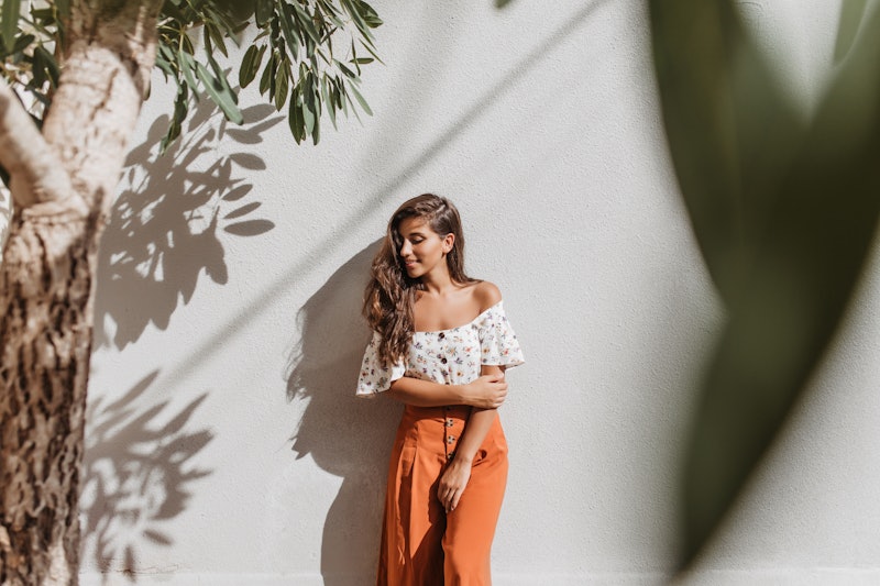 Attractive long-haired brunette woman in stylish culottes and white top posing against white wall an...