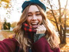 Cheerful young girl with long brown hair wearing autumn coat, walking at the park, taking a selfie