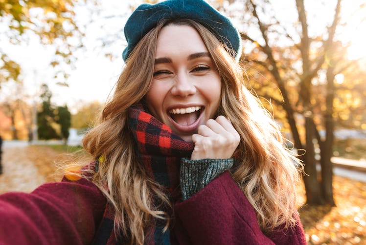 Cheerful young girl with long brown hair wearing autumn coat, walking at the park, taking a selfie