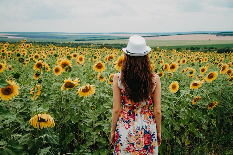 A girl in a summer dress and a white hat is standing back to frame looking at a beautiful blooming f...