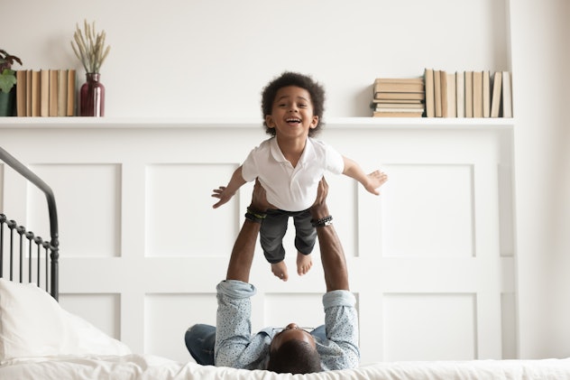 Happy funny african american kid boy flying in fathers arms looking at camera in bedroom, loving fam...