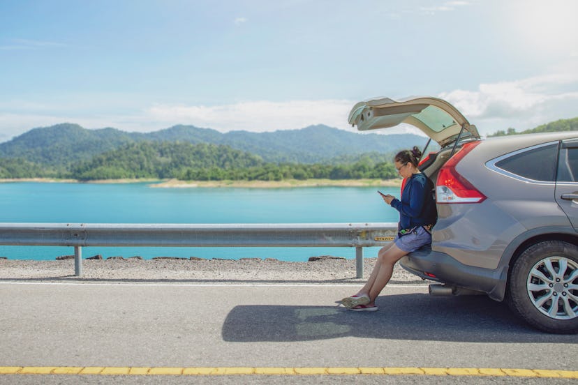 woman alone traveler on the road with sitting on hatchback car and playing smartphone with mountain ...