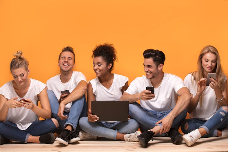 Group of young multiracial people looking at mobile phones, sitting on the floor, spending time toge...