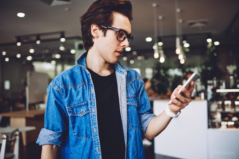 Side view of millennial hipster guy in optical spectacles for vision correction typing text for send...
