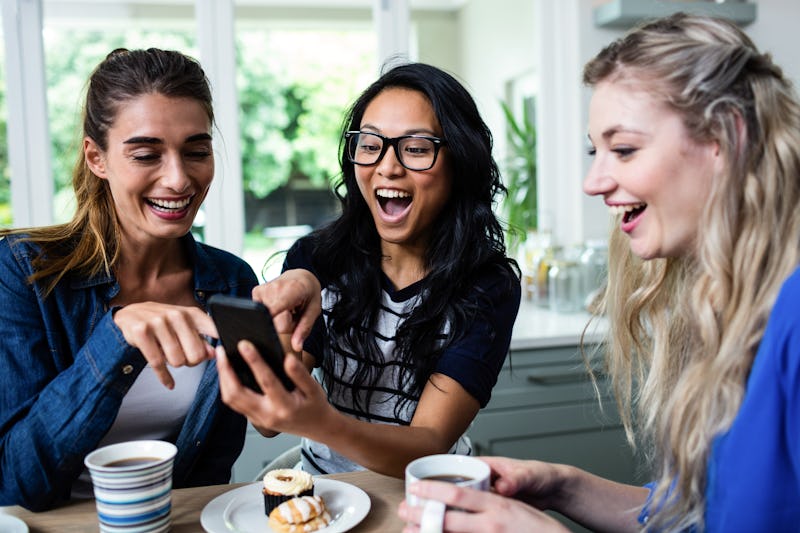 A group of friends sit around a kitchen table with coffee mugs looking at a phone.