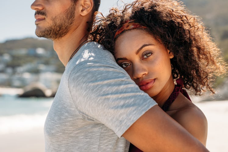 Side view of a couple in love standing outdoors. Woman holding her boyfriend from behind hugging him...
