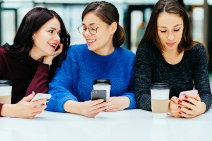 Group of three female students smiling, talking and looking in smart phone while drinking coffee fro...