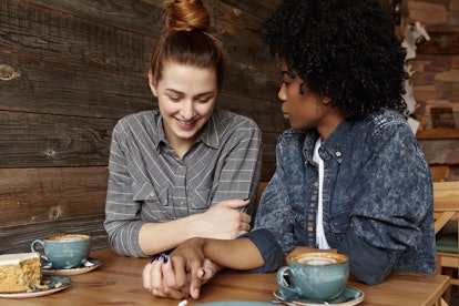 Interracial lesbian couple looking down with shy smile, holding hands during lunch at restaurant. Ha...