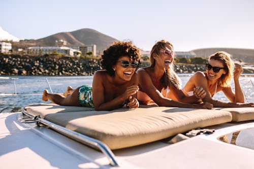 Three happy friends wearing swimsuits and sunglasses laugh while sunbathing on a yacht.