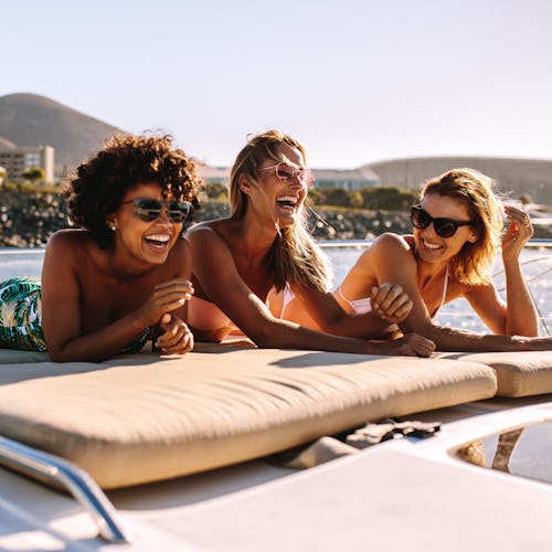 Three happy friends wearing swimsuits and sunglasses laugh while sunbathing on a yacht.