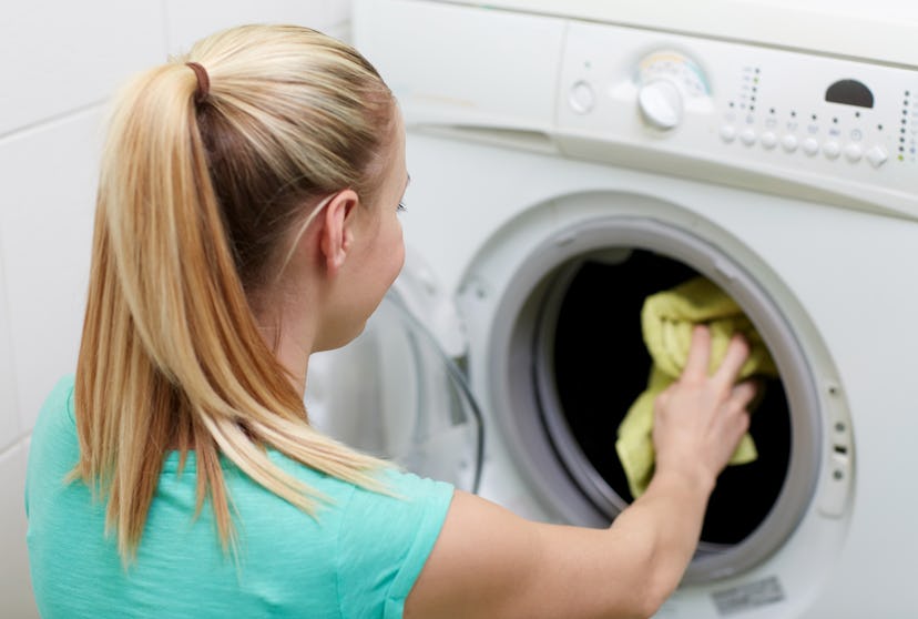 people, housework, laundry and housekeeping concept - happy woman putting laundry into washing machi...