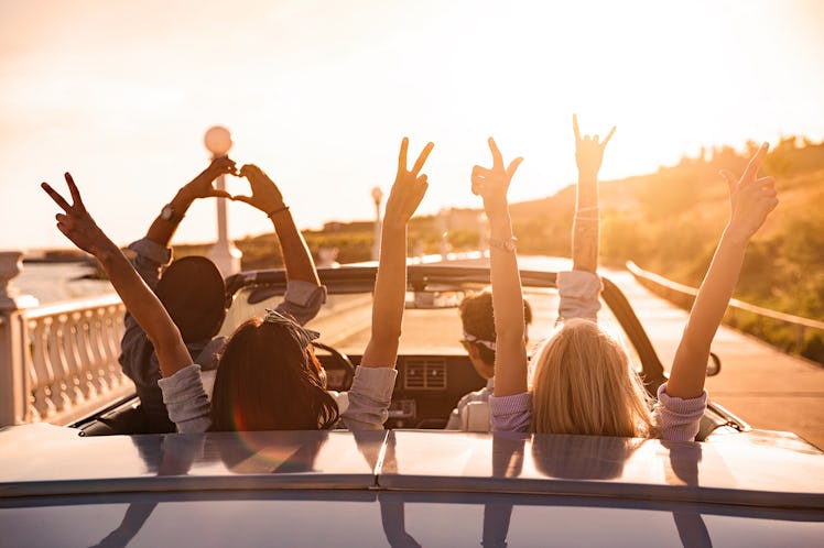 Group of happy young friends in cabriolet with raised hands driving on sunset