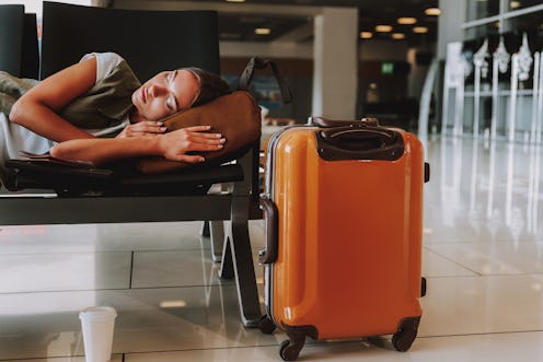 Tired young woman is waiting for plane so long that she is falling asleep on bench. Large suitcase i...