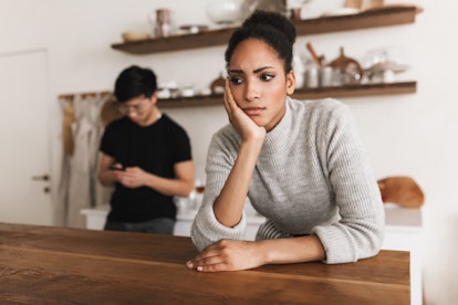 Thoughtful african american woman leaning on hand sadly looking aside with asian man using cellphonr...