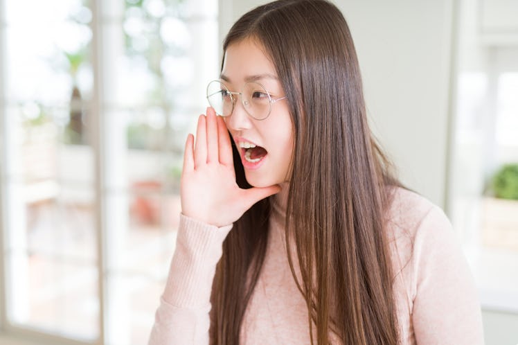 Beautiful Asian woman wearing glasses shouting and screaming loud to side with hand on mouth. Commun...