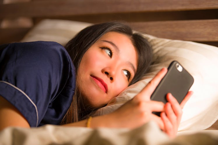 young beautiful and happy Asian Korean woman in pajamas using mobile phone social media texting with...
