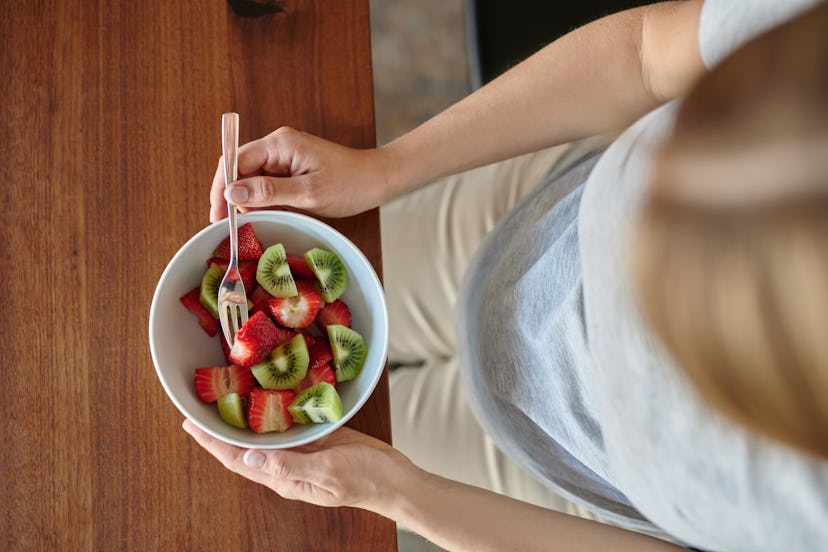 Pregnant woman eating a bowl of fruit salad with fresh strawberries seated at a dining table in a cl...