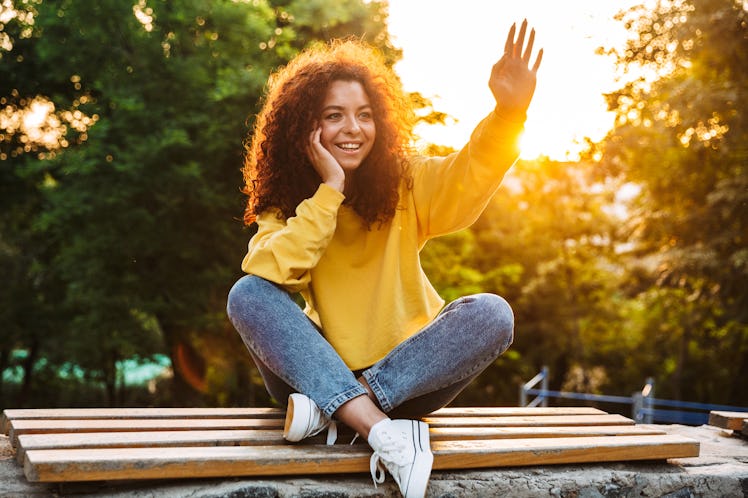 Photo of a happy cute young student curly girl sitting on bench outdoors in nature park with beautif...