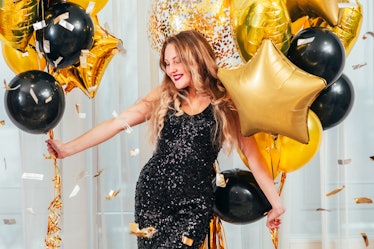 Birthday party. Happy girl in black sequin dress standing in white room with balloons. Pretty lady s...