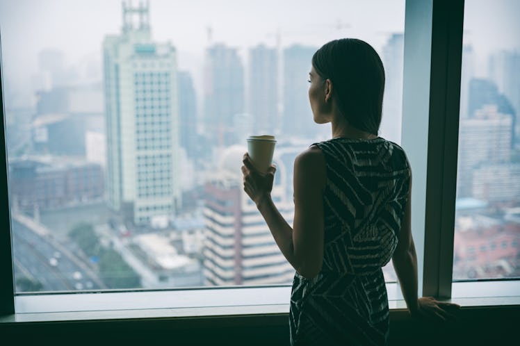 Businesswoman drinking coffee at work contemplative looking out the window of high rise skyscraper b...