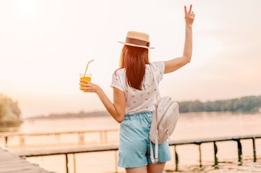 Beautiful young woman walking with orange drink on pier at sunset in summer. 