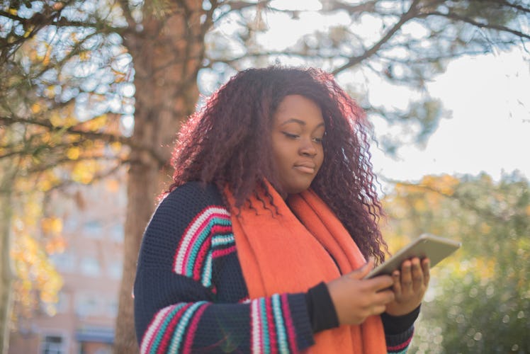 Cute smiling woman is reading pleasant text message on tablet pad while sitting in the park in warm ...