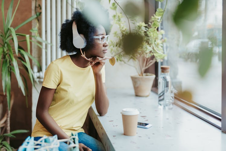 Side view of thoughtful young American girl situating near windowsill and looking through the window...