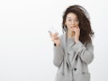 Portrait of embarrassed cute curly-haired female student in stylish grey coat and glasses, holding s...