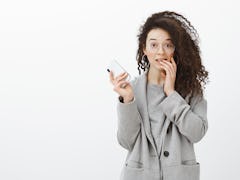 Portrait of embarrassed cute curly-haired female student in stylish grey coat and glasses, holding s...